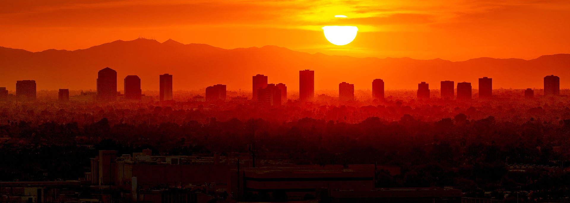 A vibrant sunset over a city skyline, featuring silhouetted buildings against a rich orange and yellow sky.